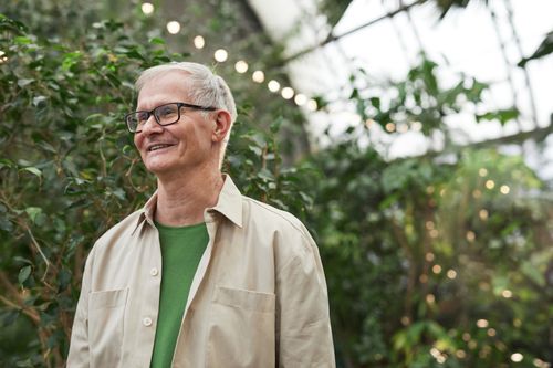 Free Elderly man smiling warmly inside a greenhouse surrounded by lush greenery. Stock Photo