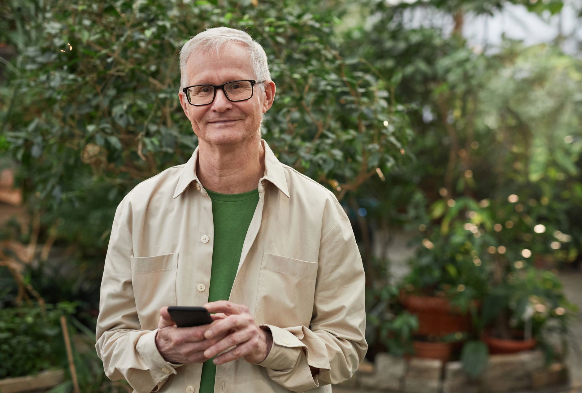 인천 어르신 스마트폰 무료 교육 2 Senior man standing in a greenhouse garden, smiling while using a smartphone. Natural sunlight.