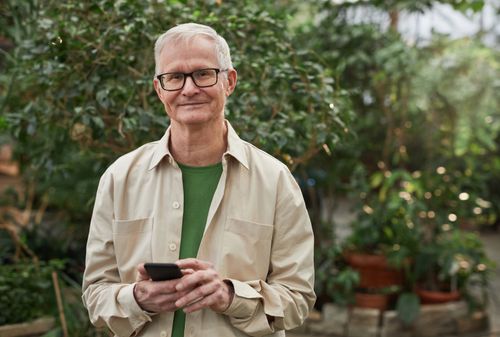 Free Senior man standing in a greenhouse garden, smiling while using a smartphone. Natural sunlight. Stock Photo