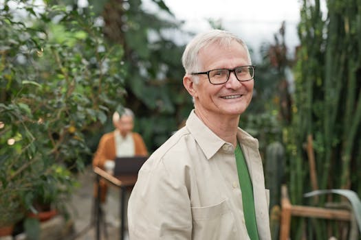 A cheerful senior man wearing glasses smiles in a vibrant greenhouse filled with greenery.