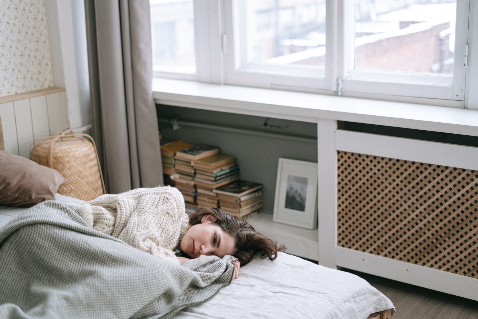 Woman in a cream knit sweater resting on a bed beside a bright window and stacked books