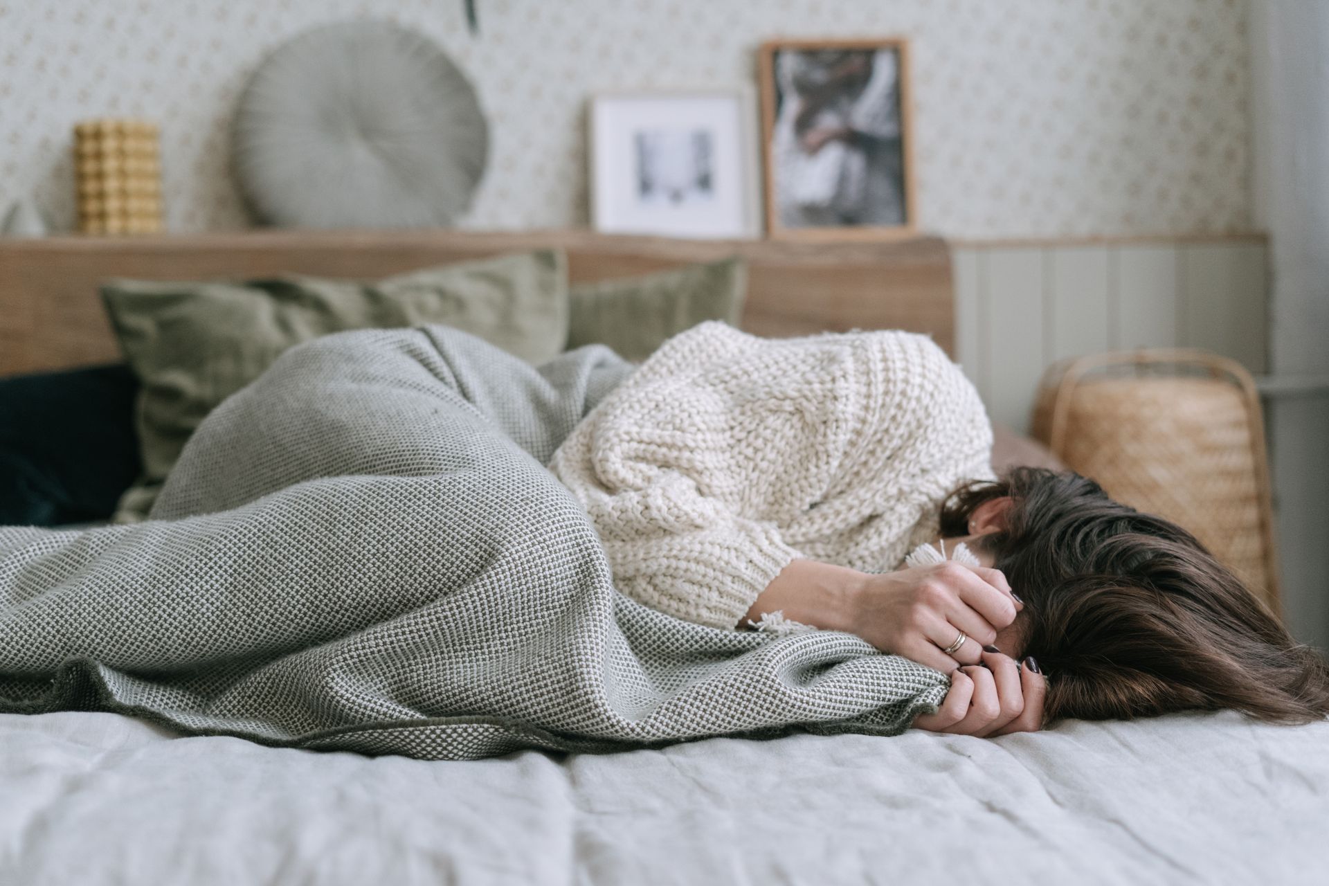 Woman wearing knitted sweater, lying on a bed indoors, wrapped in a cozy blanket for comfort.