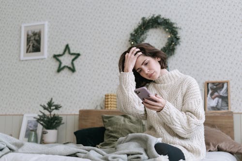 A Woman in White Knitted Sweater Sitting on Bed while Using a Smartphone