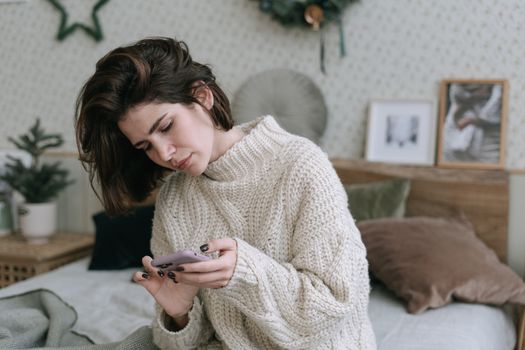 A woman in a knitted sweater using her smartphone while sitting on a bed in a cozy, decorated room.