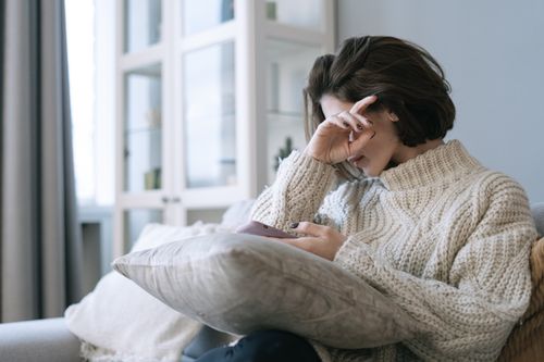 Free A woman sits indoors in a cozy knit sweater, covering her face with one hand, exuding a peaceful and introspective vibe. Stock Photo