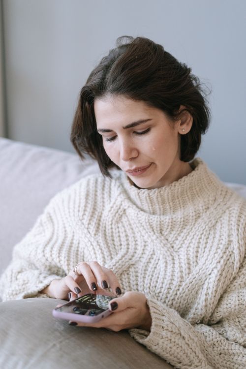 Free Pensive woman in a warm sweater using her smartphone indoors, conveying a moment of introspection. Stock Photo