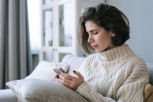 Free Woman in knitted sweater using smartphone on a cozy sofa indoors. Stock Photo