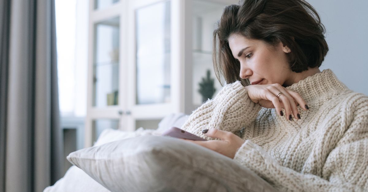 A woman in a knitted sweater using a smartphone, sitting pensively on a couch at home.