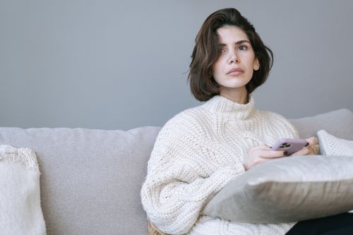 Free A thoughtful woman in a cozy sweater relaxing on a sofa while using her smartphone indoors. Stock Photo