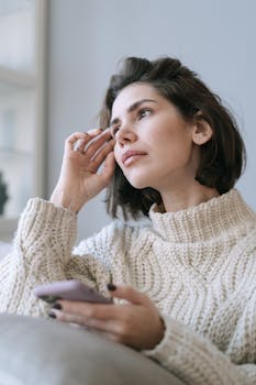 Thoughtful woman holding a smartphone indoors, wearing a knitted sweater, lost in contemplation.