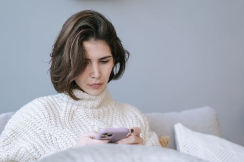 Free Woman in cozy knit sweater using a smartphone indoors, expressing a thoughtful mood. Stock Photo