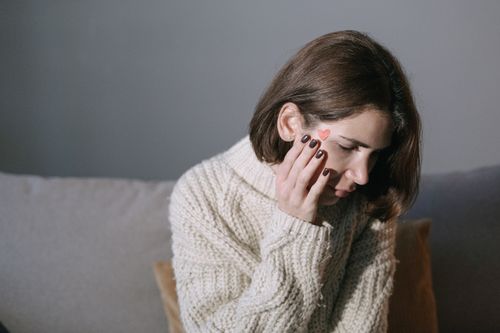 Free A woman wearing a sweater rests her hand on her face, displaying a heart sticker, in a softly lit room. Stock Photo