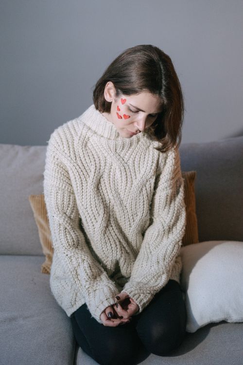 Free Woman in a knitted sweater sitting thoughtfully on a sofa with heart stickers on her face. Stock Photo