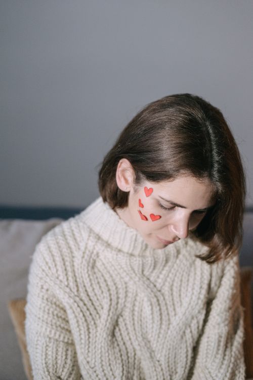 Free Woman in a knitted sweater with heart stickers on her face, looking contemplative indoors. Stock Photo