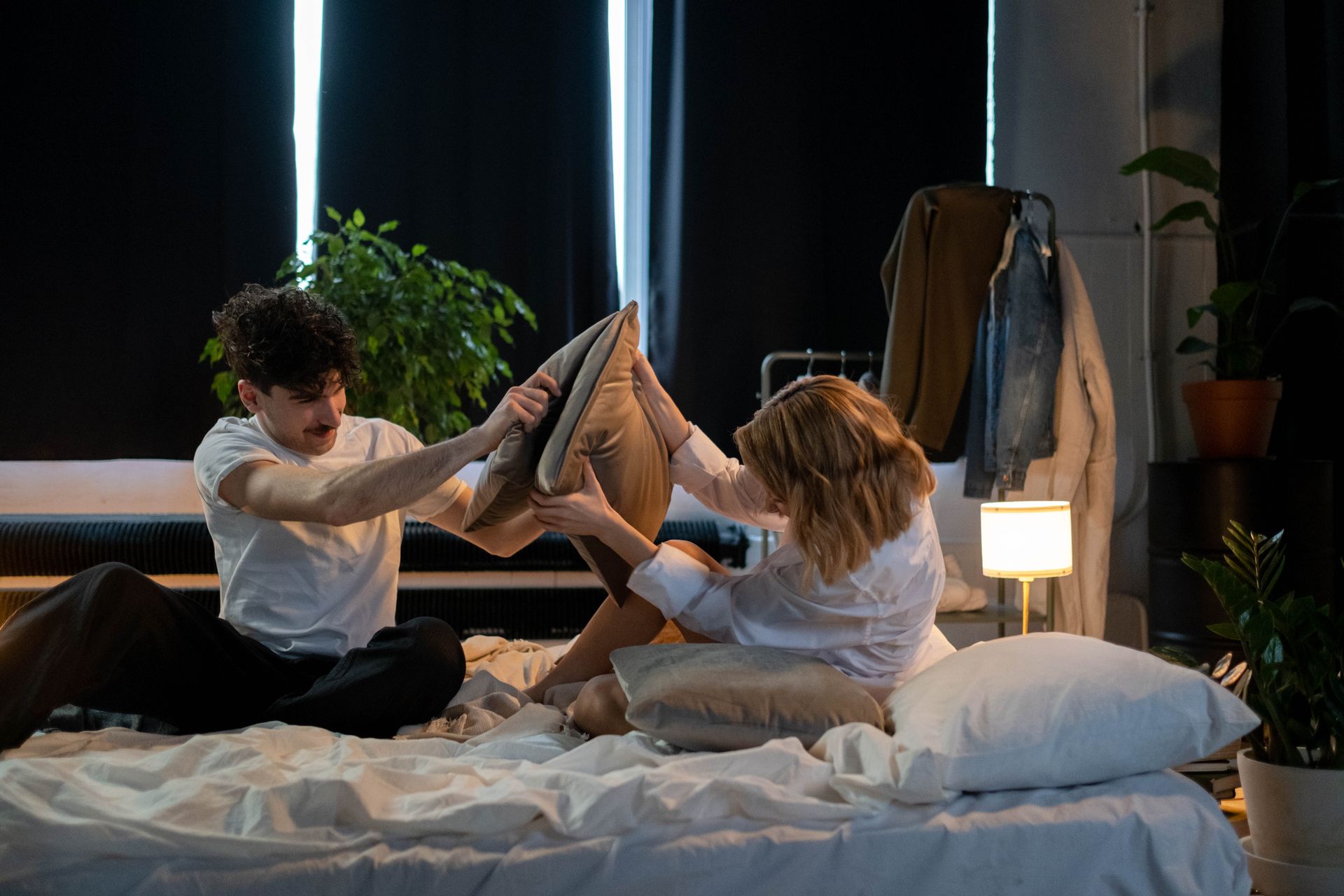 Couple enjoying a fun pillow fight in a warmly lit, modern bedroom setting.