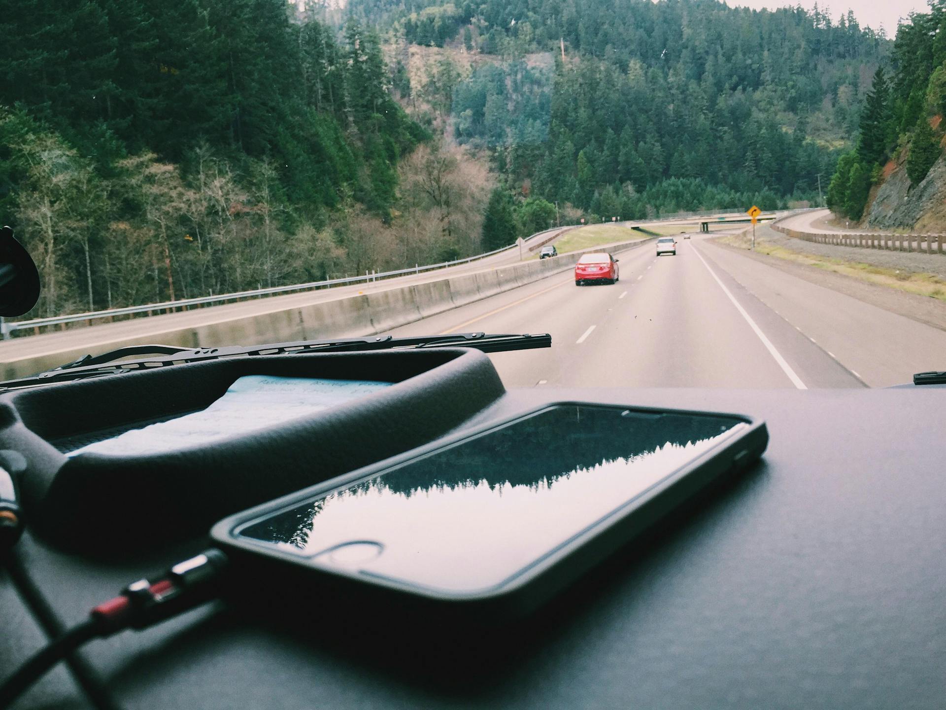 A smartphone charging on the dashboard of a car during a scenic road trip through a forested highway.