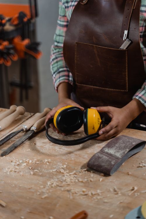 Free Artisan in leather apron holding ear protection among woodworking tools in a workshop. Stock Photo