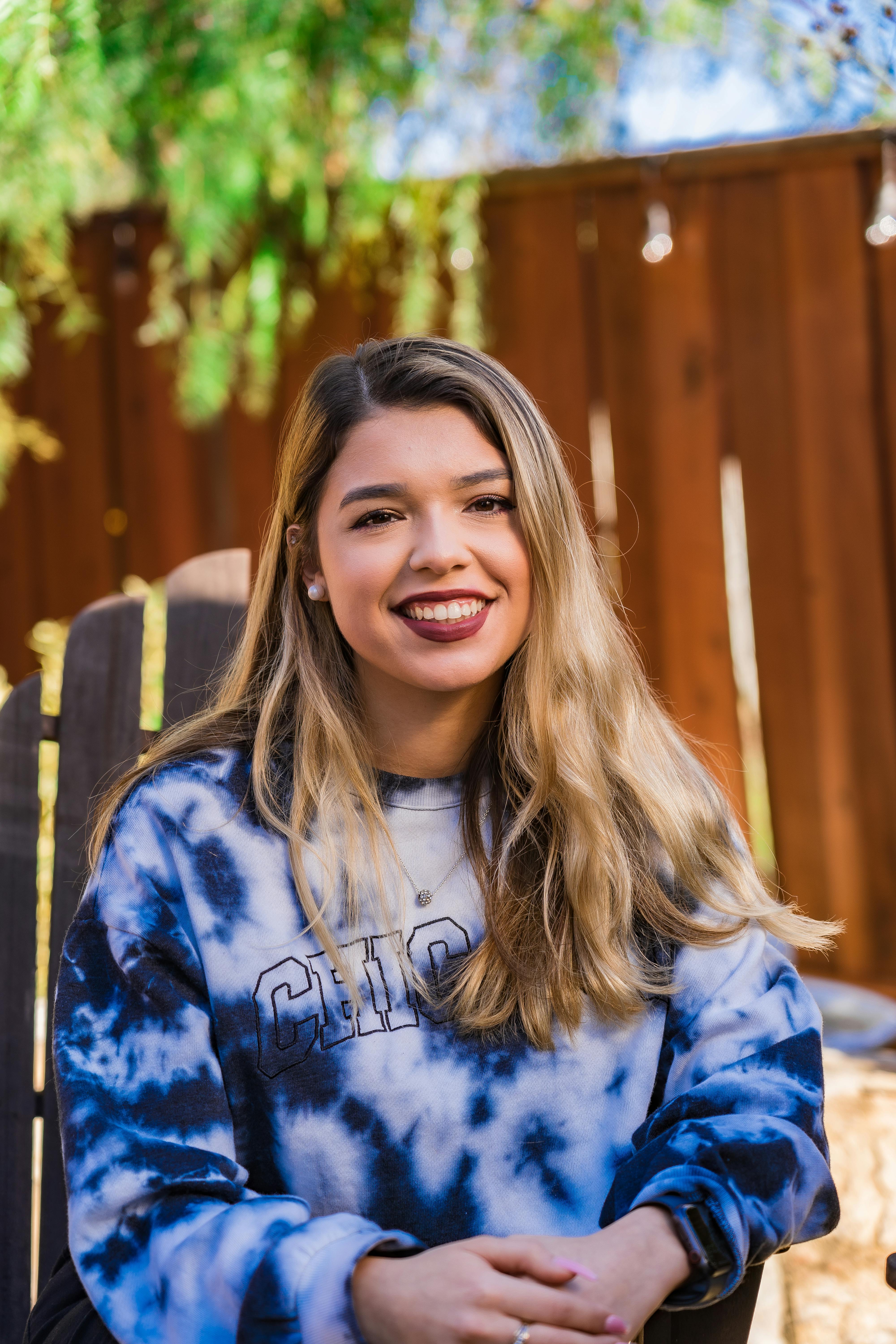 Joyful young woman with long hair wearing a stylish tie-dye sweatshirt, sitting outdoors and smiling.