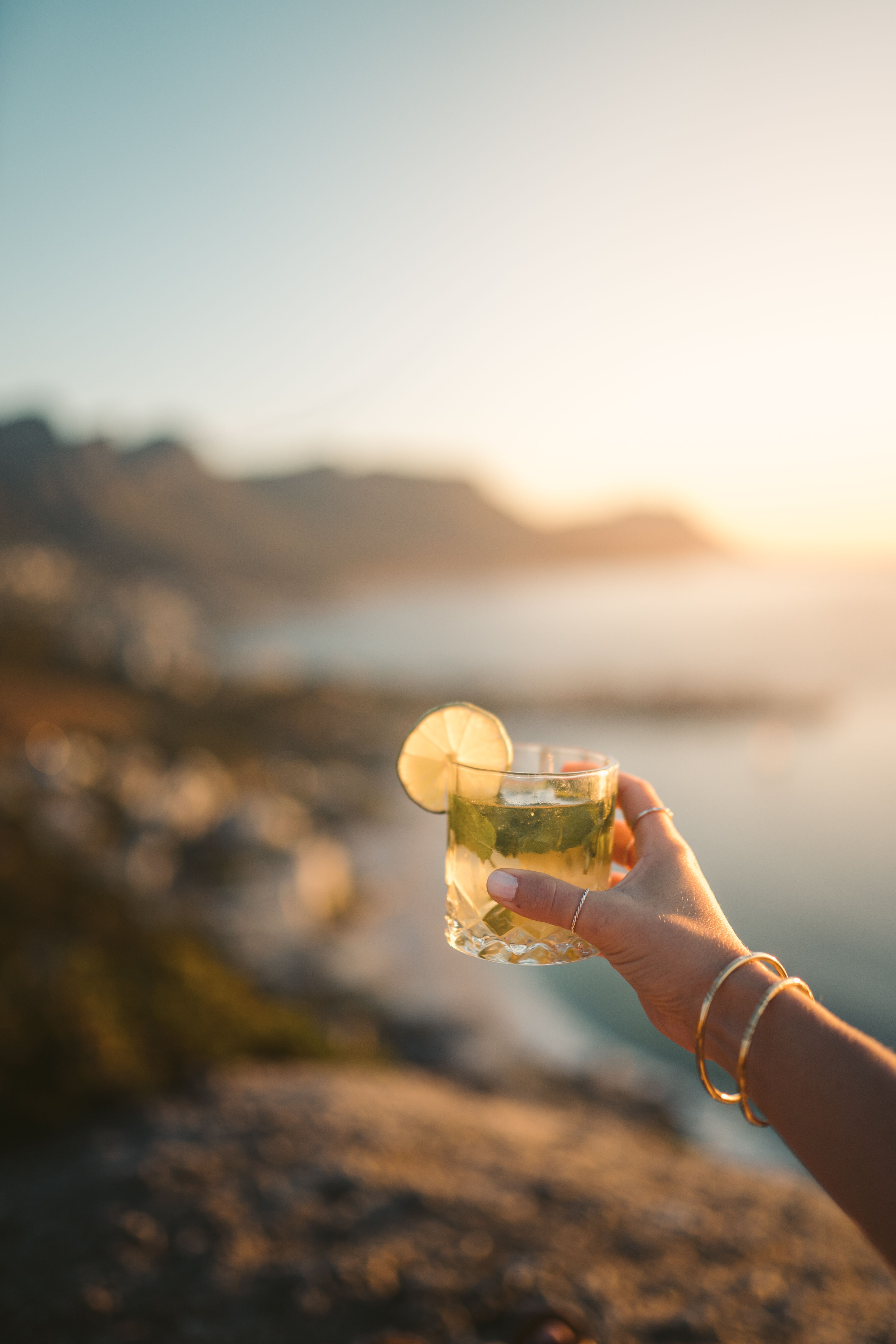 Hand holding cocktail with lime garnish against a stunning ocean sunset backdrop - The Best Happy Hours on the South Coast of Barbados