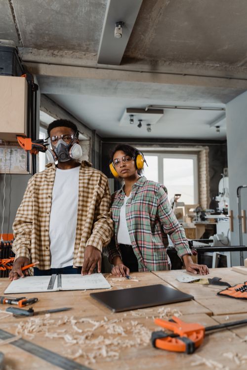 Free Two adults in a woodworking workshop with tools and protective gear, emphasizing diversity and craftsmanship. Stock Photo