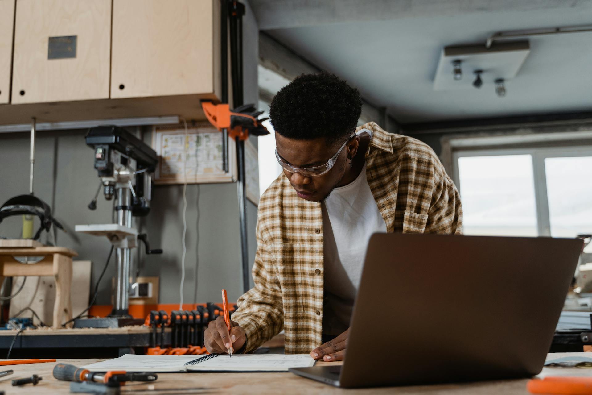 Carpenter engrossed in woodworking project with laptop and tools in a modern workshop.