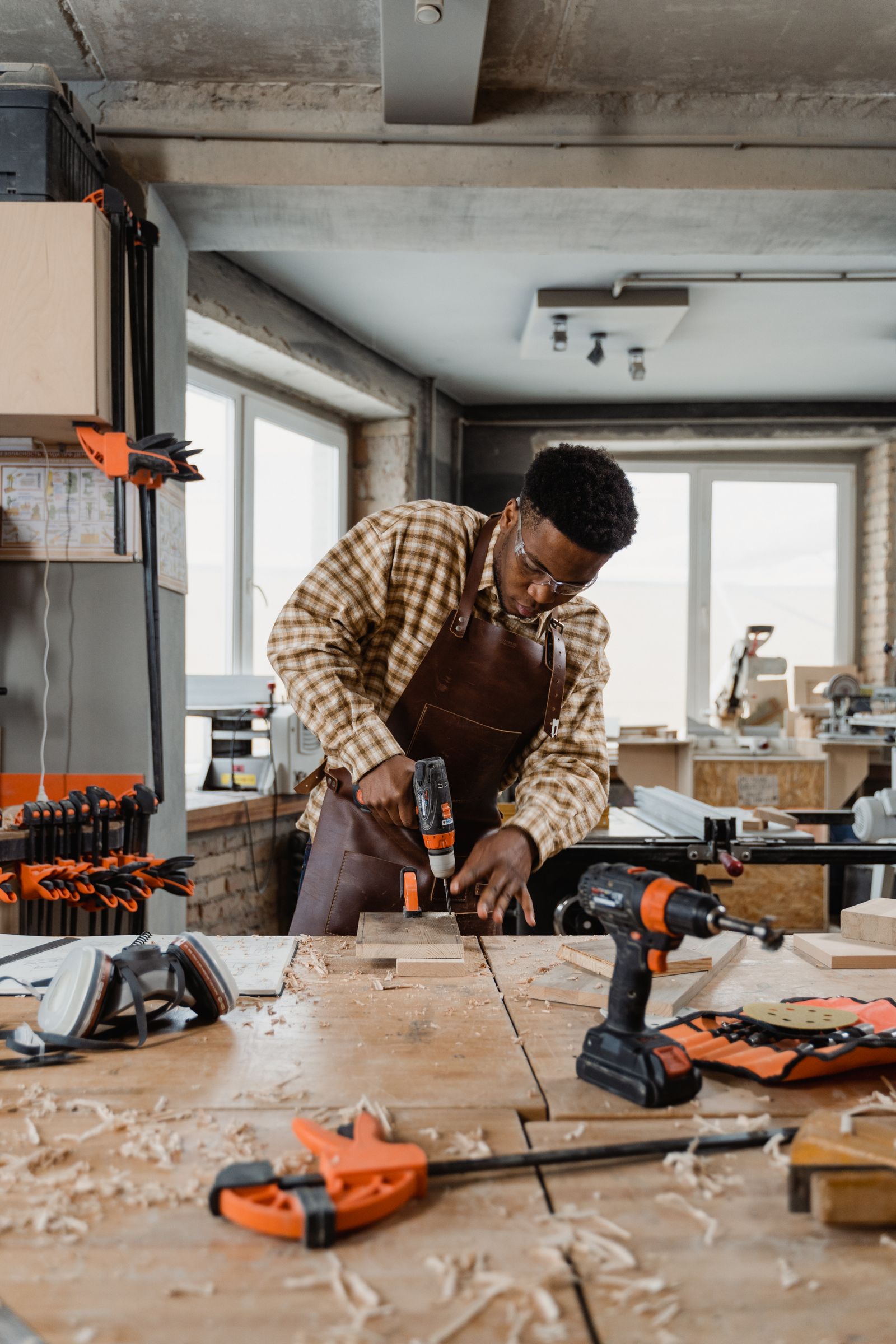 Carpenter wearing safety glasses using a power drill on wood in a bright workshop