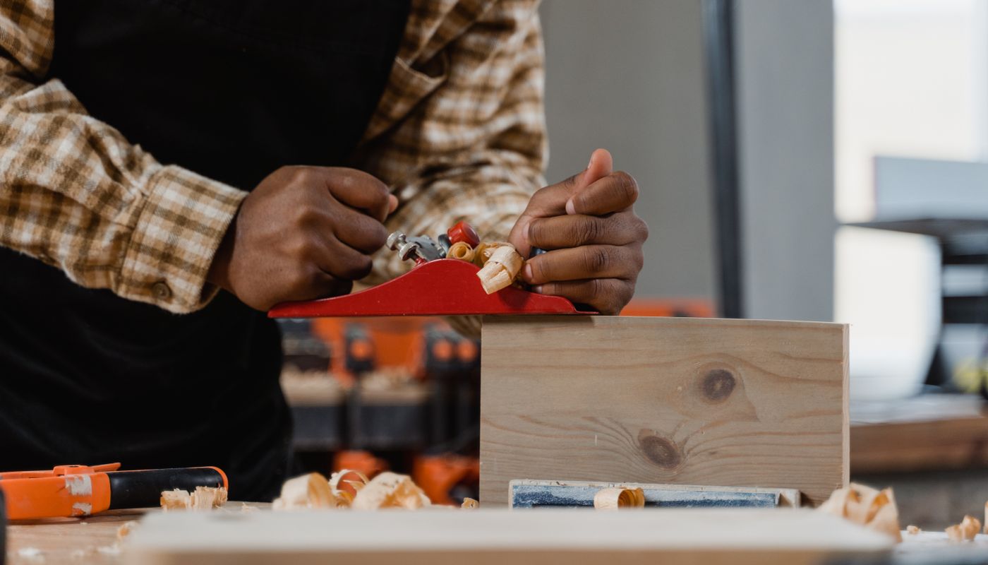 Two hands guiding a red hand plane along a pine board on a workbench, curls of shaving rising from the blade