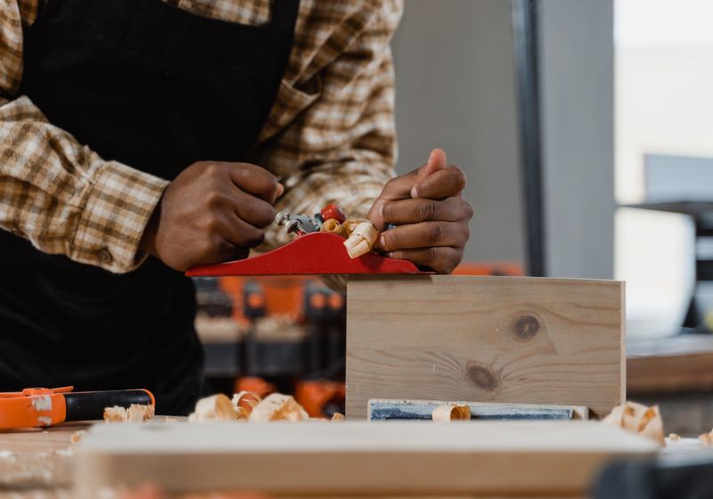 Two hands guiding a red hand plane along a pine board on a workbench, curls of shaving rising from the blade