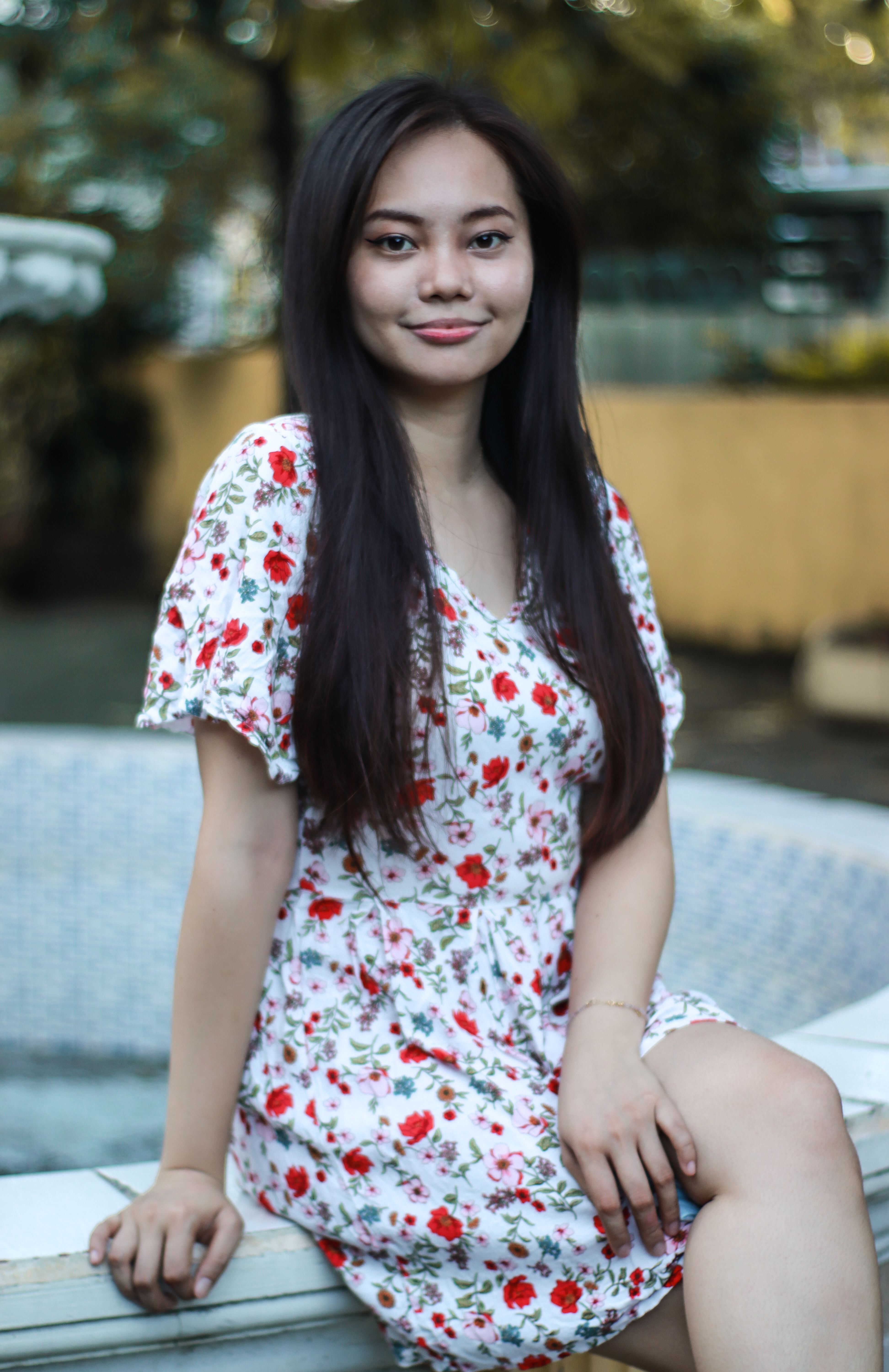 A young Asian woman wearing a floral dress sits outdoors with a gentle smile.
