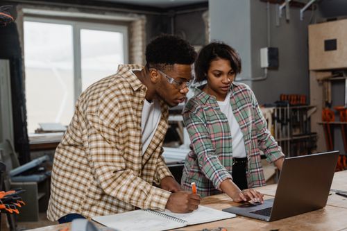 Free Two diverse young adults collaborating on a project using a laptop in a workshop environment. Stock Photo