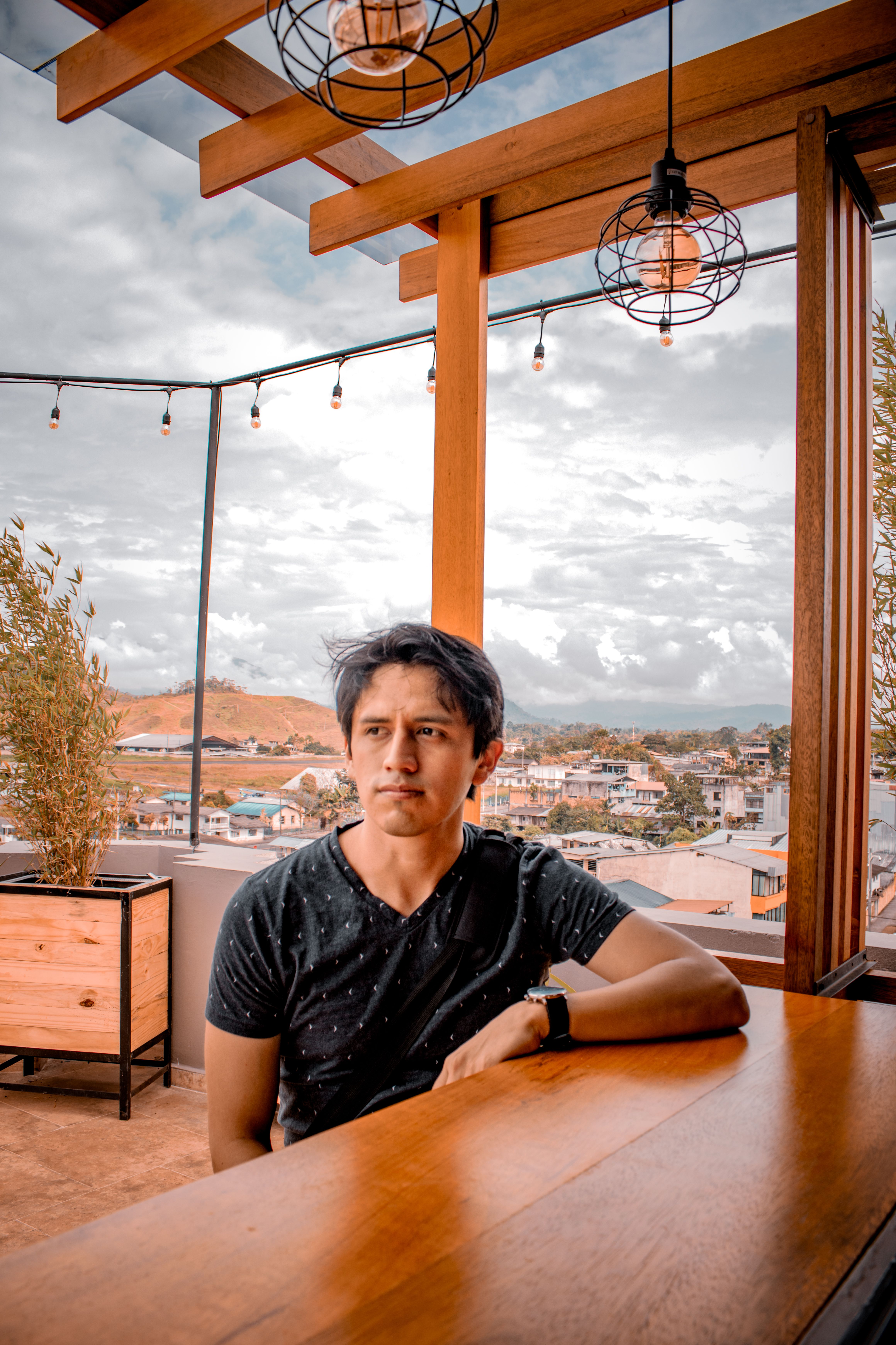 A thoughtful man sitting indoors at a rooftop bar with a scenic city view.