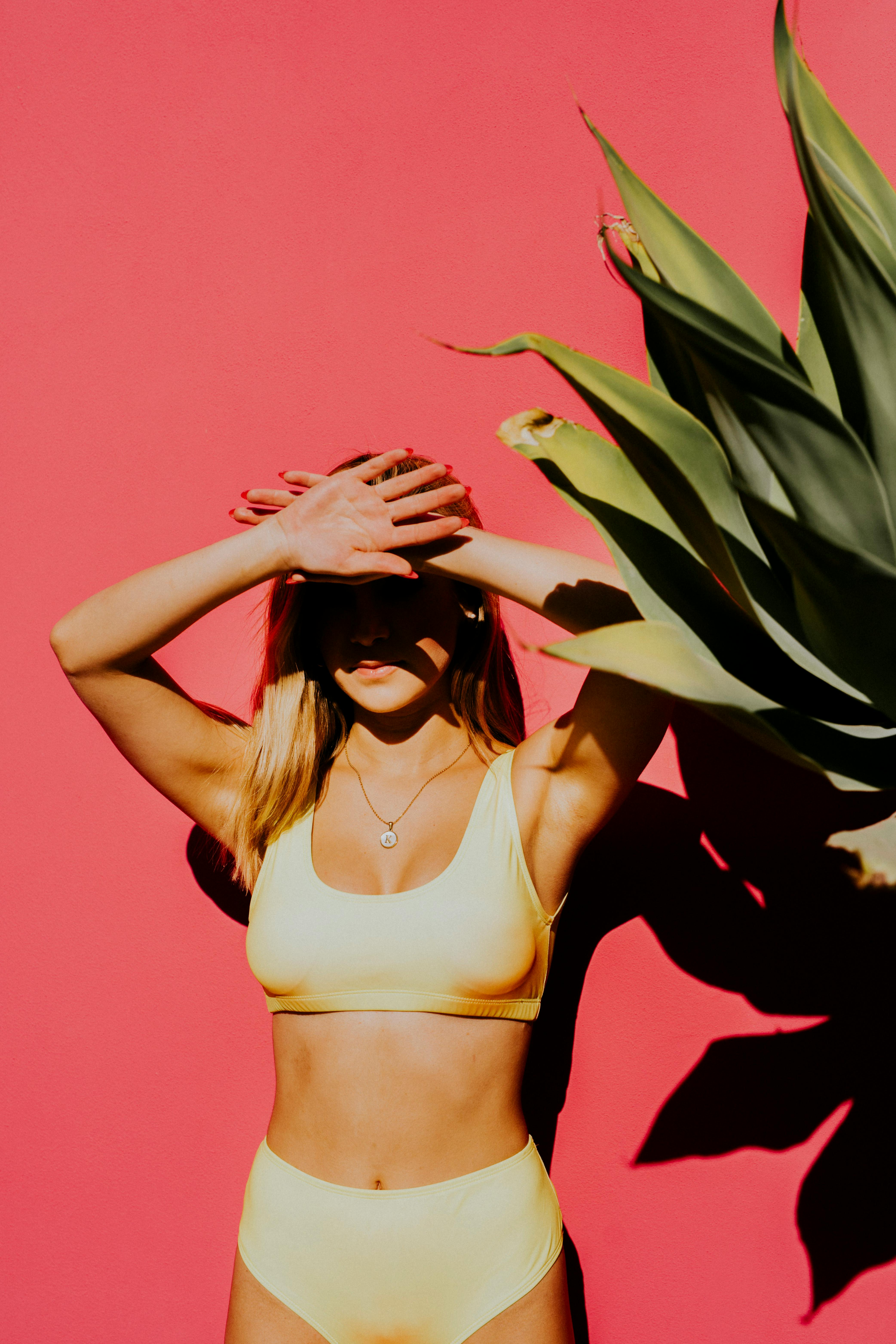 Stylish woman in yellow swimwear poses with agave shadow on a vibrant pink background.