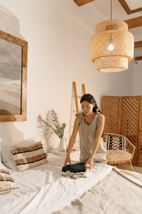 Free Asian woman folding clothes in a stylish, minimalist bedroom with warm décor and pendant lighting. Stock Photo