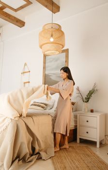 Asian woman tidying up a modern bedroom with natural decor and warm lighting.