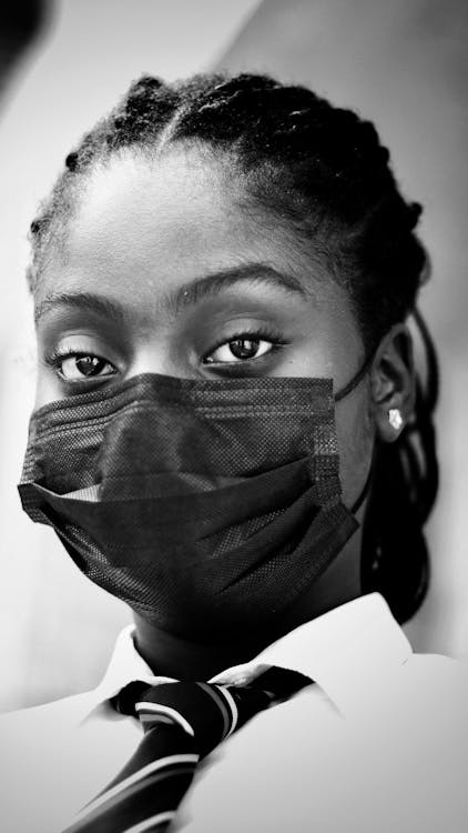 Black and white on unrecognizable young African American lady in formal outfit and medical mask looking at camera while standing near wall
