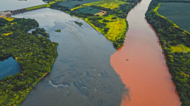Picturesque drone view of confluence of two colored rivers flowing through lush green forests and fields on sunny day in Brazil