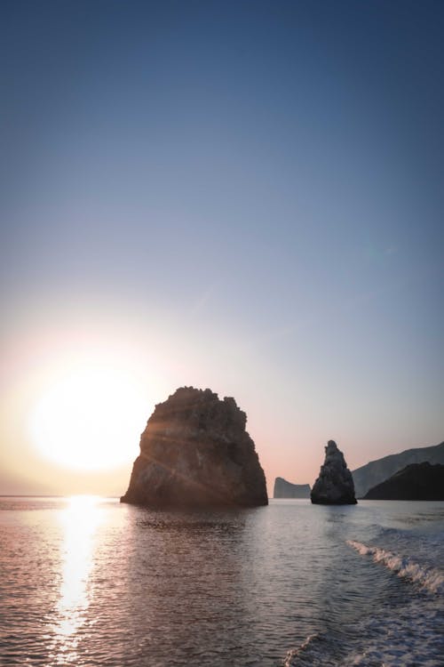 Silhouettes of rocky cliffs in wavy ocean against bright cloudless sunset sky reflecting in rippling water