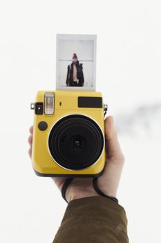 Hand holding a yellow instant camera with freshly developed photo in winter setting.