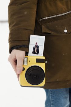 A close-up image of a yellow instant camera held by a person in a brown coat outdoors.