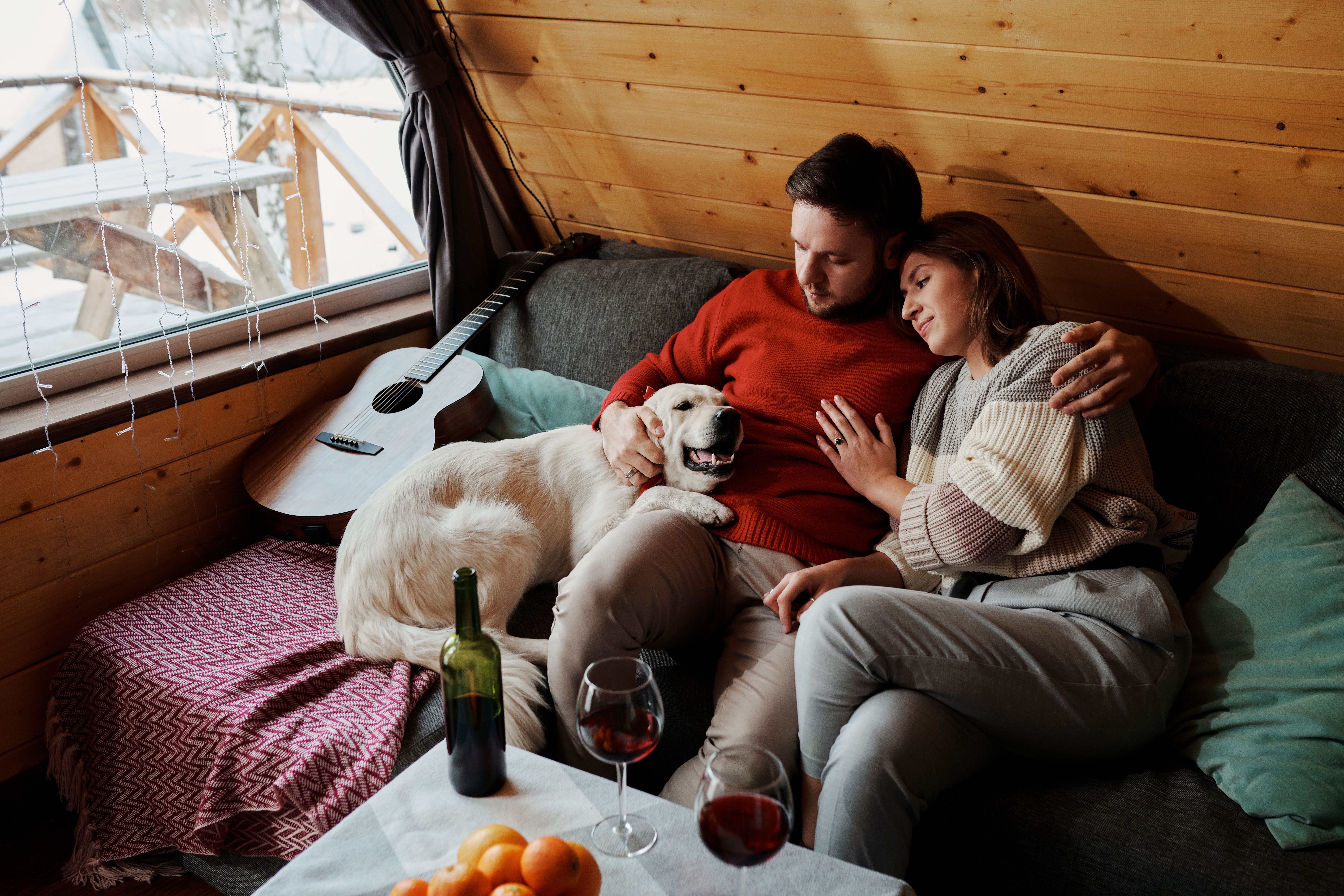 Couple relaxing with a dog in a cozy cabin, enjoying wine and warmth in a winter setting.
