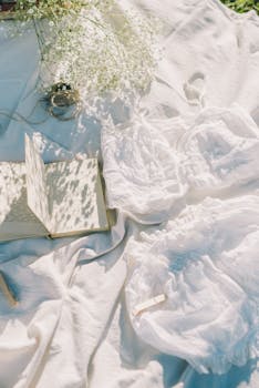 Elegant picnic setup with book, delicate flowers, and soft white textiles in sunlight.