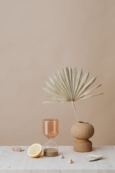 Serene minimalist composition featuring a palm leaf, hourglass, and lemon slice on a neutral background.