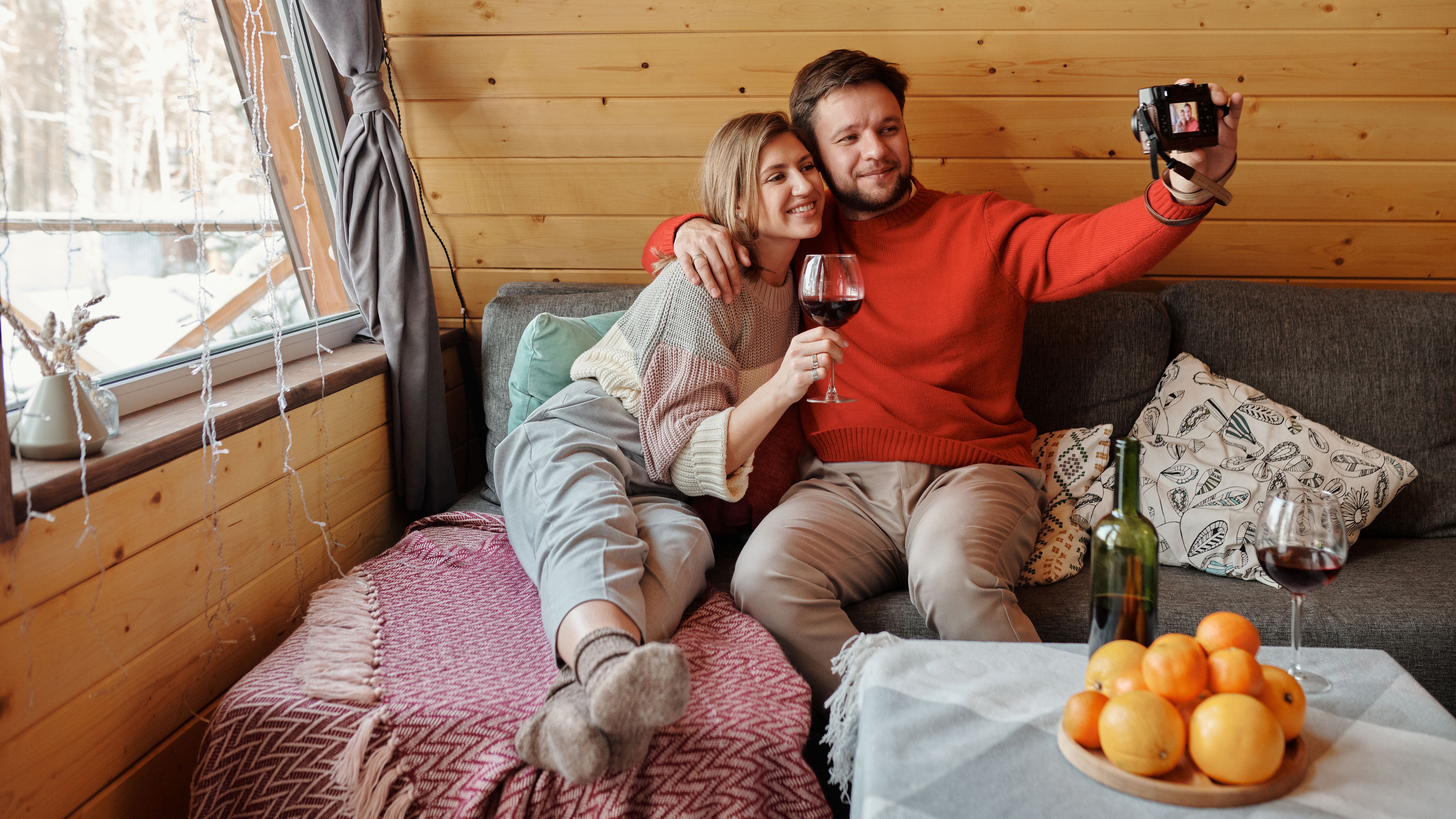 Happy couple relaxing in a wooden cabin, enjoying wine and taking selfies on a winter day.