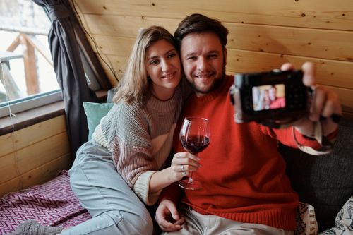 Free A happy couple takes a selfie indoors, enjoying a relaxed day with wine on the couch. Stock Photo