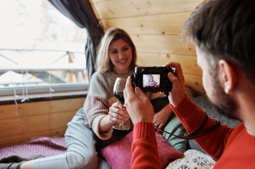 Free A couple enjoys a cozy winter day in a cabin, capturing memories with wine and a camera. Stock Photo