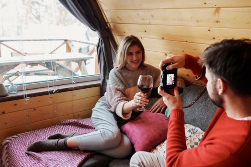 Free A couple relaxing indoors, enjoying wine and taking photos in a warm cabin atmosphere. Stock Photo