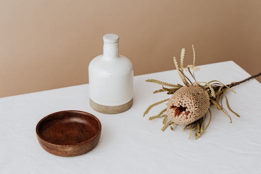 Elegant composition featuring a white bottle, wooden bowl, and dried flower on a table.