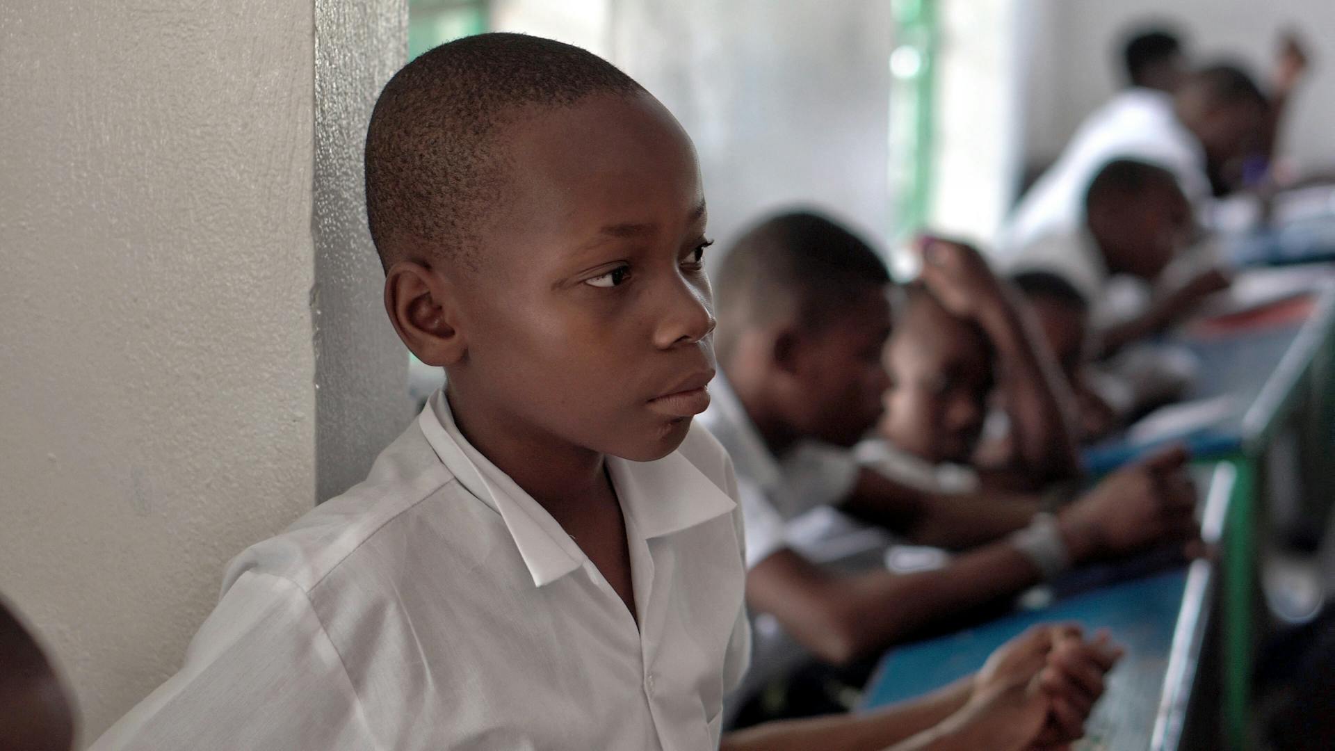 Portrait of a thoughtful young boy in a Haitian classroom, wearing a white shirt.