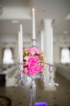 Elegant metal candelabrum with white candles decorated with composition of flowers and green leaves on blurred background of hall