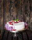 Tasty sweet cake covered with white gum paste and decorated with berries green leaves and flowers on cake plate placed on background of wooden wall